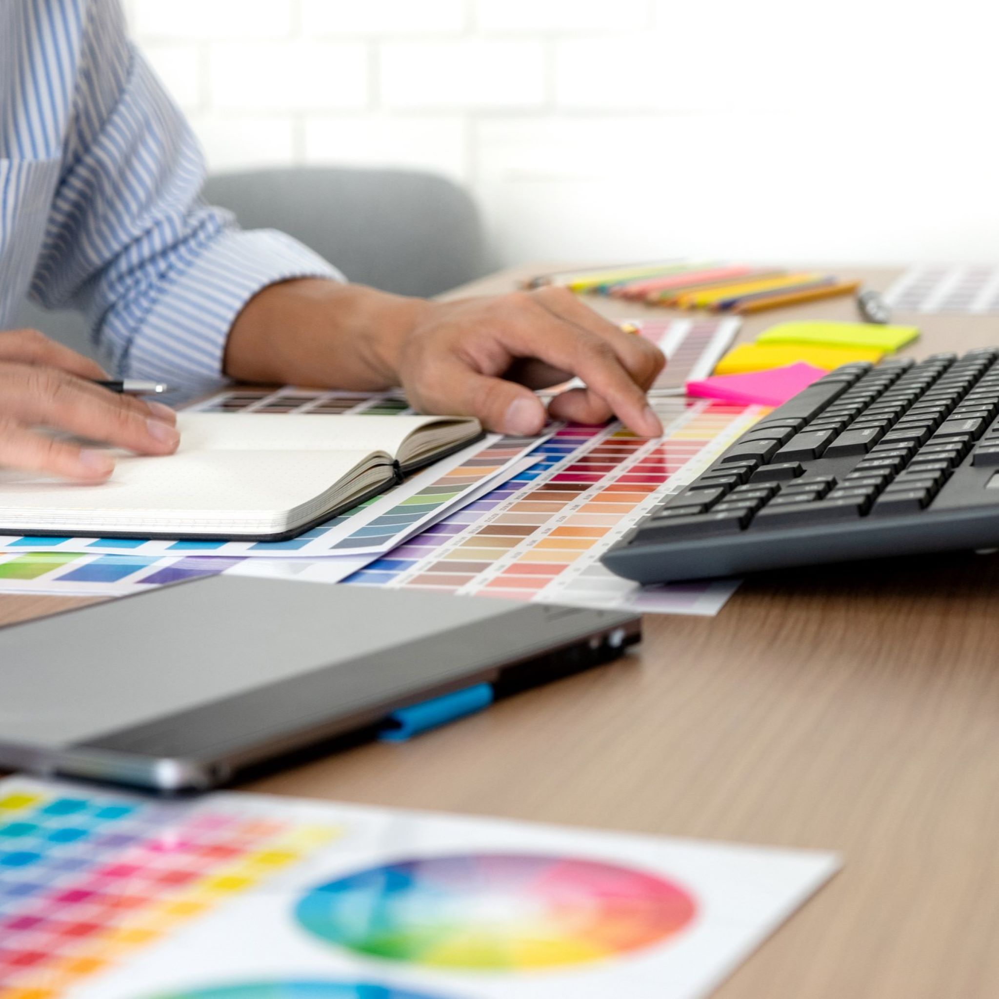 A man in a shirt sits at a desk, only his hands are visable on the desk. He has one hand on an open notebook and another pointing at a colour on a designers colour chart. A computer keyboard is on the desk in front of him and a designer's tablet is next to the keyboard.