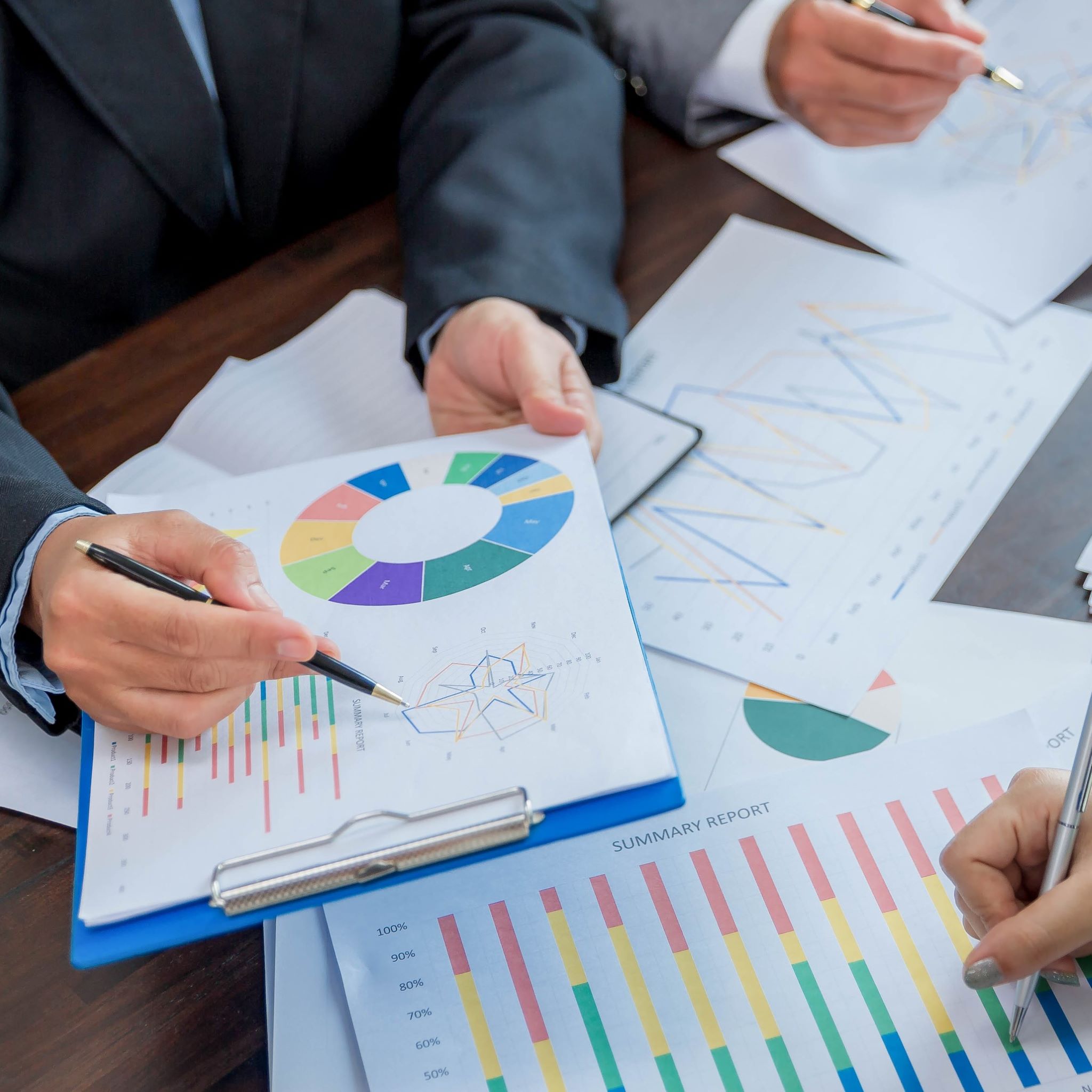 Three people sit at a table. Only their hands are visable. The all hold pens and are looking at colourful graphs and reports printed on white paper in front of them.